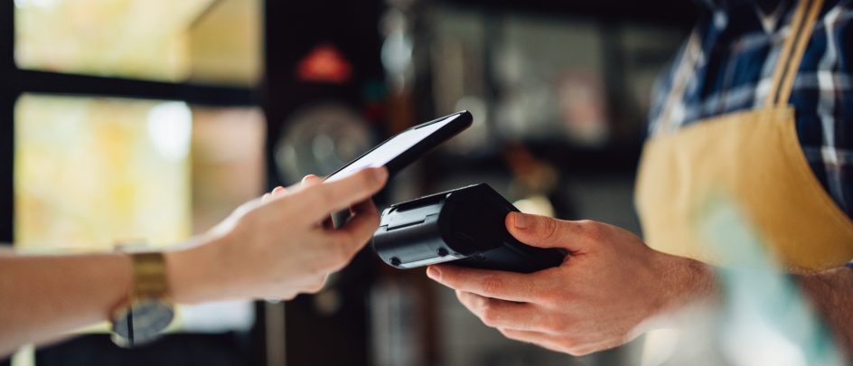 Mann med forkle holder en betalings terminal. En hånd fra venstre betaler med mobilen.