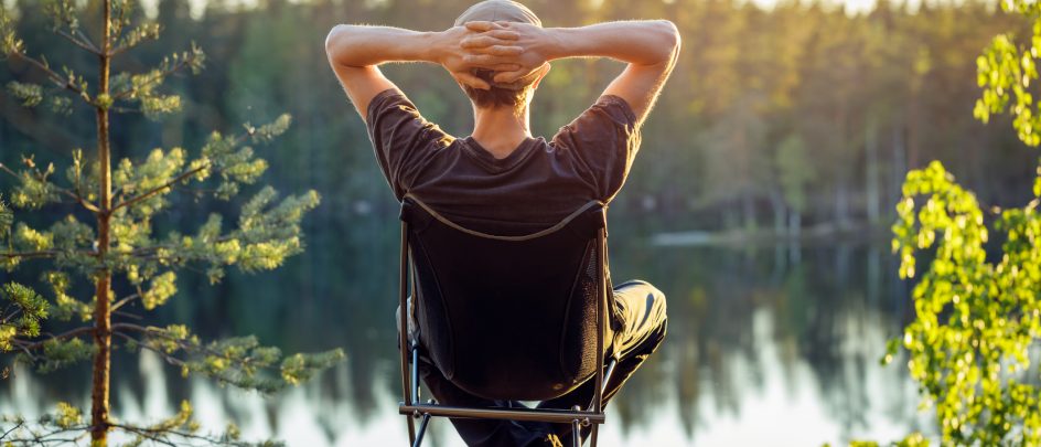 Mann som sitter i en camping stol ute i skogen og ser ut mot vannet mens solen går ned.